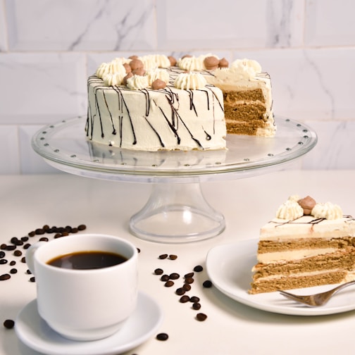 A layered cake with cream and chocolate drizzle is displayed on a glass stand. The cake is decorated with dollops of cream and small round chocolates on top. A slice of the cake is served on a plate with a fork, and a cup of black coffee sits nearby. Coffee beans are scattered on the white surface, adding contrast.