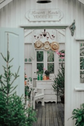 A cozy interior space with rustic decor, featuring a white wooden structure with small windows. There are potted plants on a white drawer and a wooden floor. Three straw hats hang from hooks above, under a vintage chandelier. The setting is enhanced by greenery visible through the windows, and pink flowers adding color to the scene.