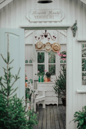 A cozy interior space with rustic decor, featuring a white wooden structure with small windows. There are potted plants on a white drawer and a wooden floor. Three straw hats hang from hooks above, under a vintage chandelier. The setting is enhanced by greenery visible through the windows, and pink flowers adding color to the scene.