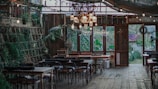 A rustic indoor setting with wooden floors and tables. The space is decorated with an assortment of chairs around several tables, surrounded by green plants and fairy lights hanging from the ceiling. A neon 'Happy Birthday' sign is visible in the background, indicating a birthday celebration is taking place. There are large windows allowing natural light to stream in, and various wrapped gifts are placed near the windows.