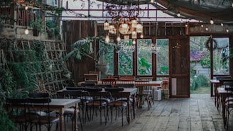 A rustic indoor setting with wooden floors and tables. The space is decorated with an assortment of chairs around several tables, surrounded by green plants and fairy lights hanging from the ceiling. A neon 'Happy Birthday' sign is visible in the background, indicating a birthday celebration is taking place. There are large windows allowing natural light to stream in, and various wrapped gifts are placed near the windows.