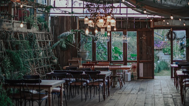 A rustic indoor setting with wooden floors and tables. The space is decorated with an assortment of chairs around several tables, surrounded by green plants and fairy lights hanging from the ceiling. A neon 'Happy Birthday' sign is visible in the background, indicating a birthday celebration is taking place. There are large windows allowing natural light to stream in, and various wrapped gifts are placed near the windows.