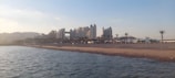 A beach scene with calm waters in the foreground and a sandy shore lined with palm trees. Several large resort buildings are visible in the background under a clear sky.