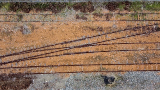 An aerial view of a railway track junction with multiple tracks diverging and converging on a gravelly surface. The ground is a mix of reddish-brown earth with patches of greenery and dry grass.