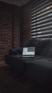 A dimly lit room features a dark gray sofa against an exposed brick wall. A laptop displaying charts and data is open on the sofa. Light filters through partially open horizontal blinds, casting shadows across the room.
