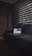 A dimly lit room features a dark gray sofa against an exposed brick wall. A laptop displaying charts and data is open on the sofa. Light filters through partially open horizontal blinds, casting shadows across the room.