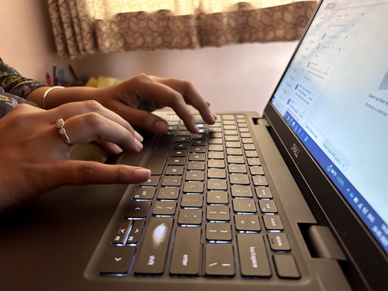 Hands typing on a laptop keyboard with a portion of the screen showing a web page. The background includes a curtain and some stationery items.
