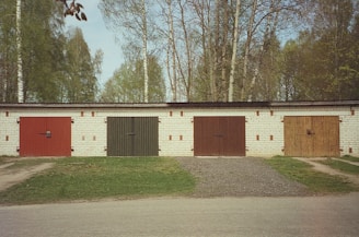 A row of four garage doors with different colors on a brick building, surrounded by a natural setting with trees in the background. The doors are red, green, brown, and wooden. The scene is serene, with a paved path in front and patches of grass.