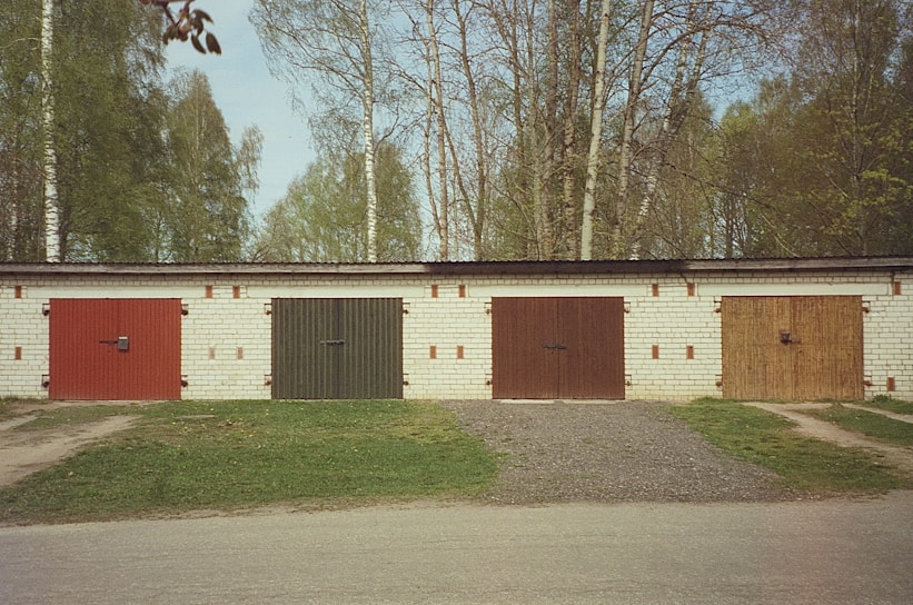 A row of four garage doors with different colors on a brick building, surrounded by a natural setting with trees in the background. The doors are red, green, brown, and wooden. The scene is serene, with a paved path in front and patches of grass.