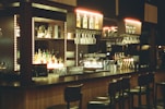 A bar setup with high stools arranged in front. The bar counter is wooden, and the back wall contains illuminated shelves holding various bottles of liquor and hanging racks of glasses. There are beer taps, a coffee machine, and other bar equipment on the counter. The lighting is soft and warm, adding to the cozy ambiance.