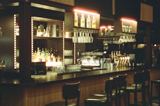 A bar setup with high stools arranged in front. The bar counter is wooden, and the back wall contains illuminated shelves holding various bottles of liquor and hanging racks of glasses. There are beer taps, a coffee machine, and other bar equipment on the counter. The lighting is soft and warm, adding to the cozy ambiance.