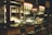 A bar setup with high stools arranged in front. The bar counter is wooden, and the back wall contains illuminated shelves holding various bottles of liquor and hanging racks of glasses. There are beer taps, a coffee machine, and other bar equipment on the counter. The lighting is soft and warm, adding to the cozy ambiance.