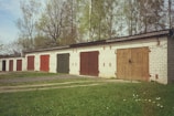 A row of garage units with colored doors stands amidst a backdrop of tall trees. The garages are made of brick, and each door is painted in different shades such as red, green, and brown. The scene is set during what appears to be a clear day, with green grass and a few scattered daisies in the foreground.