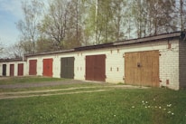 A row of garage units with colored doors stands amidst a backdrop of tall trees. The garages are made of brick, and each door is painted in different shades such as red, green, and brown. The scene is set during what appears to be a clear day, with green grass and a few scattered daisies in the foreground.