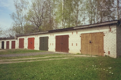 A row of garage units with colored doors stands amidst a backdrop of tall trees. The garages are made of brick, and each door is painted in different shades such as red, green, and brown. The scene is set during what appears to be a clear day, with green grass and a few scattered daisies in the foreground.