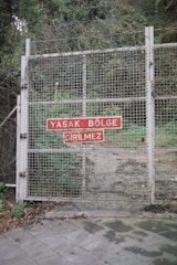 A metal gate is secured with a mesh wire fence, reinforced with barbed wire. A red sign with white text in Turkish is affixed to the gate. Vegetation is visible through and around the fence, indicating a restricted area.