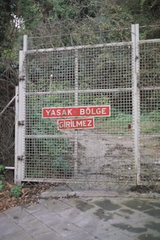 A metal gate is secured with a mesh wire fence, reinforced with barbed wire. A red sign with white text in Turkish is affixed to the gate. Vegetation is visible through and around the fence, indicating a restricted area.