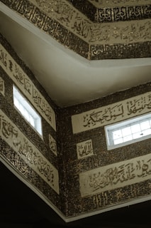 Ornate Arabic calligraphy decorates the walls and ceilings of an architectural structure. The intricate lettering is surrounded by elaborate patterns and illuminated by small windows that let in natural light.