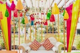 An intricately decorated sofa with colorful fabric tassels and garlands hanging above and around it. The sofa features ornate designs with two vibrant red and yellow patterned cushions. Bright vertical drapes of pink, yellow, and green fabric frame the scene, with floral and bead accents enhancing the aesthetic.