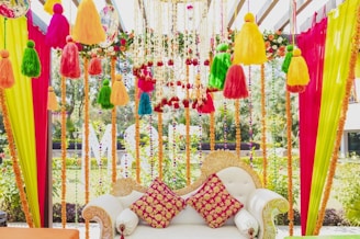 An intricately decorated sofa with colorful fabric tassels and garlands hanging above and around it. The sofa features ornate designs with two vibrant red and yellow patterned cushions. Bright vertical drapes of pink, yellow, and green fabric frame the scene, with floral and bead accents enhancing the aesthetic.