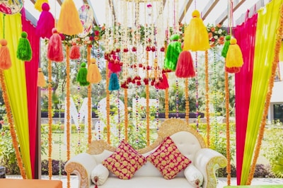 An intricately decorated sofa with colorful fabric tassels and garlands hanging above and around it. The sofa features ornate designs with two vibrant red and yellow patterned cushions. Bright vertical drapes of pink, yellow, and green fabric frame the scene, with floral and bead accents enhancing the aesthetic.
