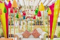 An intricately decorated sofa with colorful fabric tassels and garlands hanging above and around it. The sofa features ornate designs with two vibrant red and yellow patterned cushions. Bright vertical drapes of pink, yellow, and green fabric frame the scene, with floral and bead accents enhancing the aesthetic.