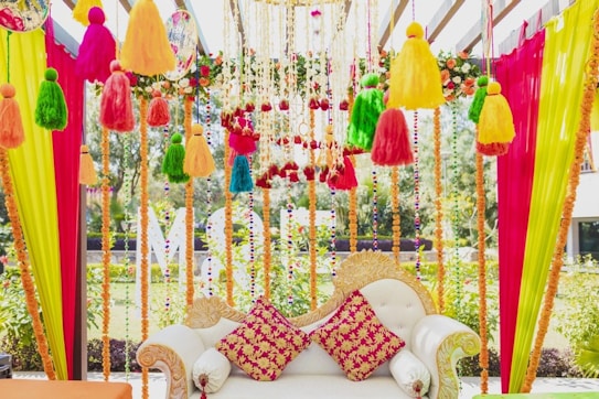 An intricately decorated sofa with colorful fabric tassels and garlands hanging above and around it. The sofa features ornate designs with two vibrant red and yellow patterned cushions. Bright vertical drapes of pink, yellow, and green fabric frame the scene, with floral and bead accents enhancing the aesthetic.