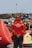 A person wearing a red jacket with the text 'LES SAUVETEURS EN MER SNSM' stands with hands clasped behind their back in front of numerous rescue boats. The setting appears to be a maritime rescue operation, with a large buoy to the left and a rocky shoreline in the background. Several rescue and safety equipment items are visible.