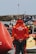 A person wearing a red jacket with the text 'LES SAUVETEURS EN MER SNSM' stands with hands clasped behind their back in front of numerous rescue boats. The setting appears to be a maritime rescue operation, with a large buoy to the left and a rocky shoreline in the background. Several rescue and safety equipment items are visible.