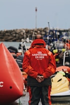 A person wearing a red jacket with the text 'LES SAUVETEURS EN MER SNSM' stands with hands clasped behind their back in front of numerous rescue boats. The setting appears to be a maritime rescue operation, with a large buoy to the left and a rocky shoreline in the background. Several rescue and safety equipment items are visible.
