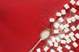 A collection of white sugar cubes scattered across a vibrant red surface. A vintage silver spoon filled with granulated sugar is placed among the cubes, adding a touch of elegance. Some granules of sugar are also sprinkled around the area, creating a textured contrast against the background.