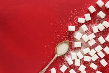 A collection of white sugar cubes scattered across a vibrant red surface. A vintage silver spoon filled with granulated sugar is placed among the cubes, adding a touch of elegance. Some granules of sugar are also sprinkled around the area, creating a textured contrast against the background.