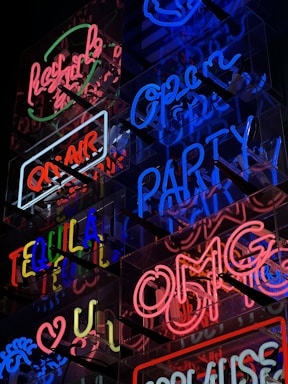 A vibrant display of neon signs featuring various phrases in bright colors such as 'OPEN', 'PARTY', 'TEQUILA', and 'OMG'. The signs are arranged on a reflective surface, creating a visually striking and lively effect.