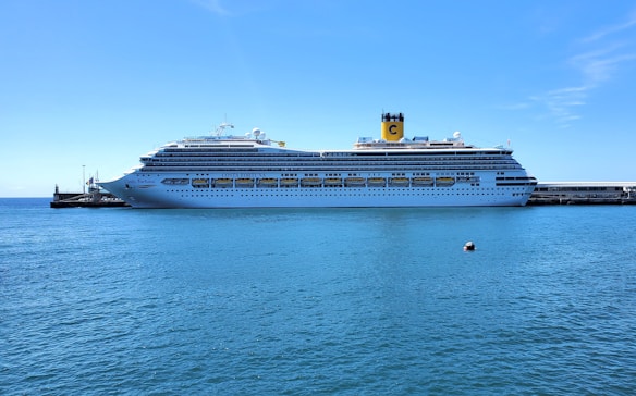 A large cruise ship docked at a pier, with a calm blue sea and clear sky in the background. The ship features multiple decks and prominent yellow lifeboats along its side, and the visible name 'Costa Fortuna' on its hull.