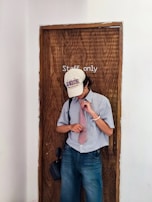 A person wearing a baseball cap with text, a striped shirt, and a red tie stands in front of a wooden door labeled 'Staff only'. They appear to be adjusting their tie and are carrying a small bag. The setting has a simple, undecorated wall.