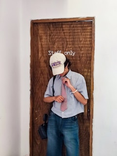 A person wearing a baseball cap with text, a striped shirt, and a red tie stands in front of a wooden door labeled 'Staff only'. They appear to be adjusting their tie and are carrying a small bag. The setting has a simple, undecorated wall.