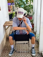 A person is seated in a chair in a relaxed pose, wearing a striped shirt, tie, and a white cap with text. They have rolled-up denim shorts and black high-top sneakers. Surrounding them are green potted plants and a checkered tile floor adds to the casual atmosphere.