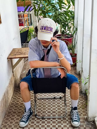 A person is seated in a chair in a relaxed pose, wearing a striped shirt, tie, and a white cap with text. They have rolled-up denim shorts and black high-top sneakers. Surrounding them are green potted plants and a checkered tile floor adds to the casual atmosphere.