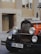An antique black and orange car is parked on a street with a background of beige buildings. The front of the car features large, round headlights and a chrome radiator grille. A European license plate with the initials 'CZ' and the number '6AN 3564' is attached to the front bumper.
