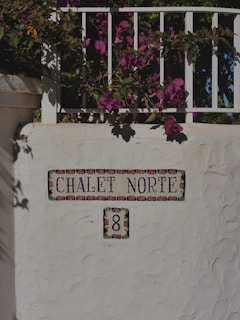 A decorative white wall with colorful floral tiles featuring the name 'Chalet Norte' and the number '8'. Above the wall, vibrant pink bougainvillea flowers drape over the railing, casting shadows on the surface.