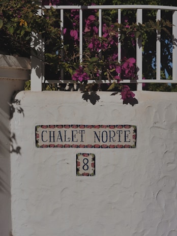 A decorative white wall with colorful floral tiles featuring the name 'Chalet Norte' and the number '8'. Above the wall, vibrant pink bougainvillea flowers drape over the railing, casting shadows on the surface.
