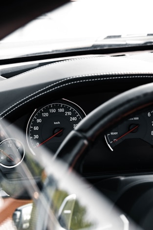 A close-up view of a car's dashboard, featuring a speedometer and tachometer. The speedometer shows a range from 0 to 260 km/h, while the tachometer provides RPM measurements. The dashboard is black with subtle stitching details, and there's a steering wheel partially visible in the foreground.