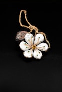 A gold chain necklace features a pendant shaped like a flower with white petals adorned with small green gems. At the center of the flower is a larger yellow gem. Behind the flower, a dark textured stone adds contrast to the composition. The background is a smooth, black surface that enhances the details of the jewelry.