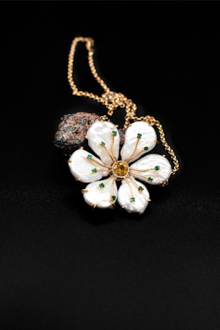 A gold chain necklace features a pendant shaped like a flower with white petals adorned with small green gems. At the center of the flower is a larger yellow gem. Behind the flower, a dark textured stone adds contrast to the composition. The background is a smooth, black surface that enhances the details of the jewelry.