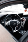 The interior of a luxury car featuring a sleek steering wheel with a chrome Mercedes-Benz logo at the center. The dashboard includes digital displays and air vents with metallic accents. A small screen is mounted on the center console, and a decorative Fanta air freshener hangs from the rear-view mirror.