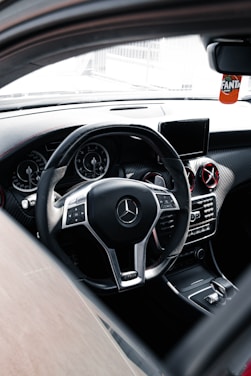 The interior of a luxury car featuring a sleek steering wheel with a chrome Mercedes-Benz logo at the center. The dashboard includes digital displays and air vents with metallic accents. A small screen is mounted on the center console, and a decorative Fanta air freshener hangs from the rear-view mirror.