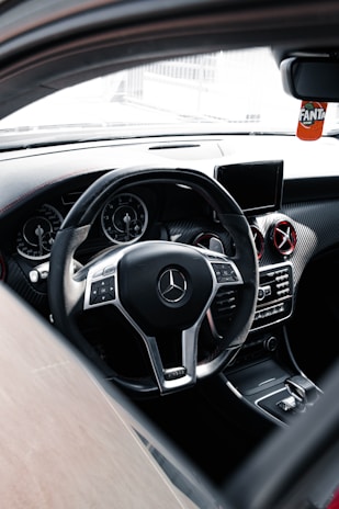 The interior of a luxury car featuring a sleek steering wheel with a chrome Mercedes-Benz logo at the center. The dashboard includes digital displays and air vents with metallic accents. A small screen is mounted on the center console, and a decorative Fanta air freshener hangs from the rear-view mirror.