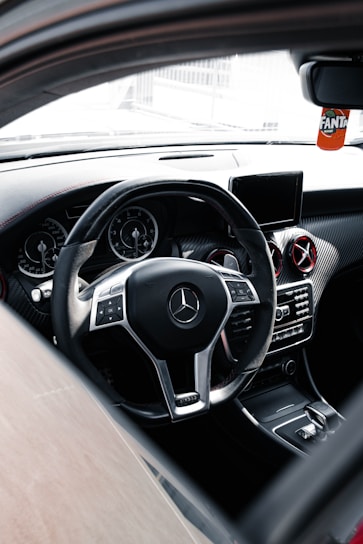 The interior of a luxury car featuring a sleek steering wheel with a chrome Mercedes-Benz logo at the center. The dashboard includes digital displays and air vents with metallic accents. A small screen is mounted on the center console, and a decorative Fanta air freshener hangs from the rear-view mirror.