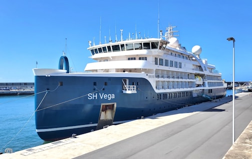 A large cruise ship named 'SH Vega' is docked at a port. The ship has multiple decks with numerous windows and satellite domes, and its hull is painted in a dark blue color. The sky is clear and sunny, illuminating the white structures of the ship. The pier is mostly empty, with a few lampposts visible.