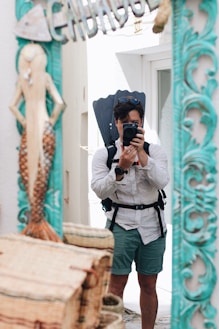 A person wearing a white shirt and green shorts is taking a photograph of themselves in a mirror. They have a backpack and a pair of diving fins attached, and sunglasses are perched on their head. The mirror is decorated with a carved mermaid and teal accents. Wicker baskets are visible in the foreground.
